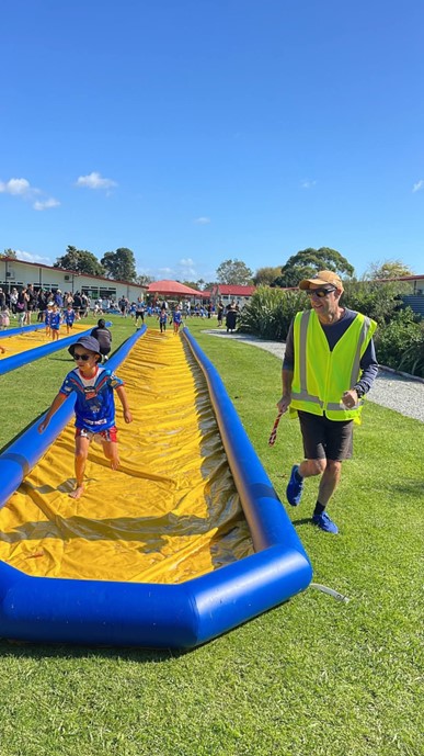 Figure 2 - Kaiti School student Te Ahiwera and GVRC Manager Tom Anderson achieving success at the Weetbix Try-athalon in April. Navigating the slippery slide! Tom is standing beside the slide and Te Ahiwera is running on the slide. The slide is on the grass and it is a very sunny day.