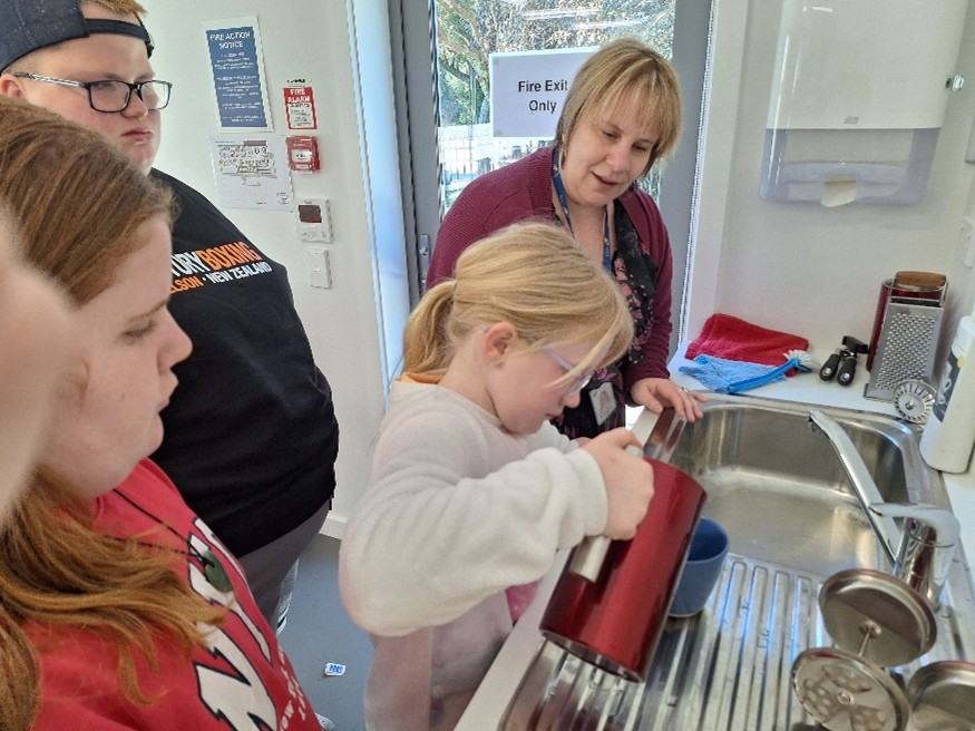 Figure 1 A akonga is standing at a bench pouring water from a jug with two other akonga watching and the Orientation Mobility instructor