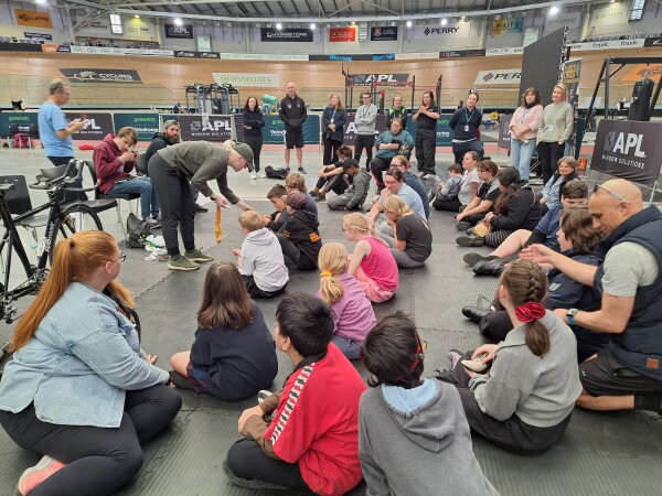 A group of BLENNZ ākonga, parents and other adults supporting the day. Some are sitting on the floor and some are standing. They are listening to a paralympic cyclist talk about what it is like to race on a tandem bike. There is a bike leaning on a chair in front of them.