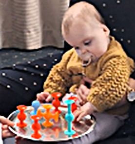 Eliza is wearing a textured mustard-coloured sweater and is seated on a dark cushioned chair. Eliza is interacting with a round metallic tray that holds several brightly coloured suction toys in orange, red, and blue. The toys are arranged upright on the tray, and an adult hand is visible holding the edge of the tray. The background includes a curtain and part of the chair’s armrest.