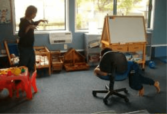 A child in a classroom with their body across a chair with wheels, pushing the chair with their legs and 'chasing' their teacher.