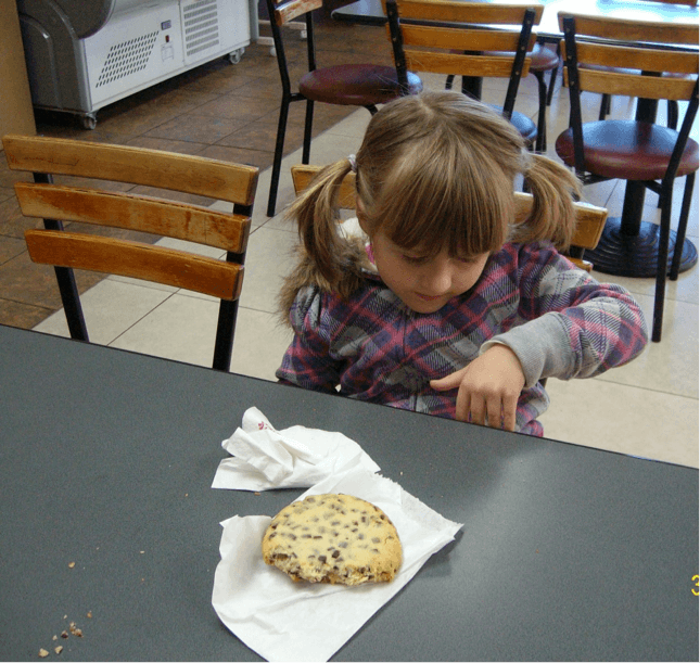 Child sitting at a table in a food court with a biscuit on the table, her left hand resting against the edge of the table.