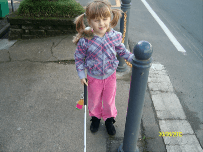 Child standing to the right of bollard beside a road crossing with cane in her right hand, trailing the chain that connects the bollards.