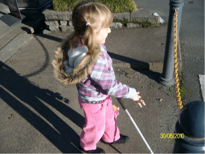 Child walking toward road crossing with cane in her left hand and right hand out in front of her.