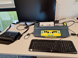 A display on a desk of a setup featuring a black computer monitor, a black keyboard, a blue and yellow Braille Mountbatten machine with paper loaded, and a black Braille display device. The arrangement highlights assistive technology tools used for Braille reading and writing alongside standard computer equipment.