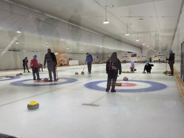 A indoor curling rink with multiple players engaged in a game, wearing helmets and winter clothing. The image features curling stones on ice sheets marked with concentric circles in blue, white, and red.