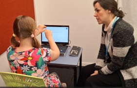 A learner is sitting on a chair with a desk in front of her. On the desk is a laptop computer. An adult is watching her while she is doing a eye tracking assessment. They are in a room.