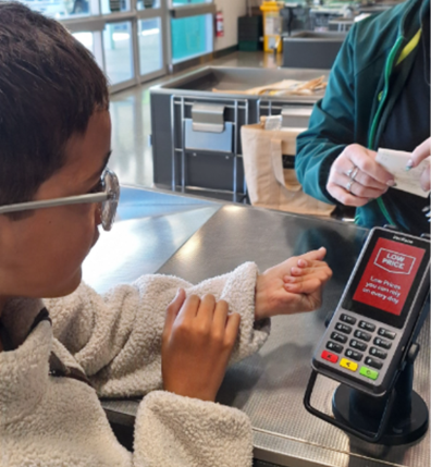A ākonga is standing at a checkout at the supermarket paying for food items using a eftpos machine and receiving a receipt from the adult behind the checkout.