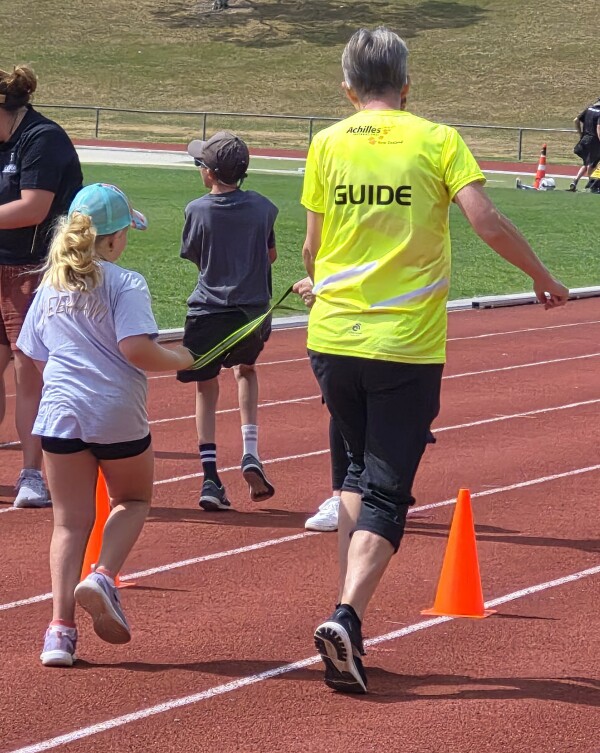 An ākonga is holding a tethering strap and running with a adult guide on a running track. There is some orange cones on the ground. There is other people in the background. It is a very sunny day.
