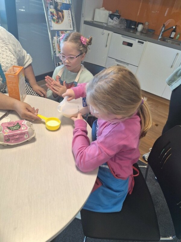 Two girls are sitting at a round table with an adult. One girl is pouring milk into a yellow measuring cup which is being held by the adult. The other girl looks on. There is a kitchen bench in the background.