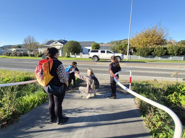 Figure 2 - There is 2 adults and 2 students on a path beside the road. One of the adults is teaching one of  the students how to cross the road.