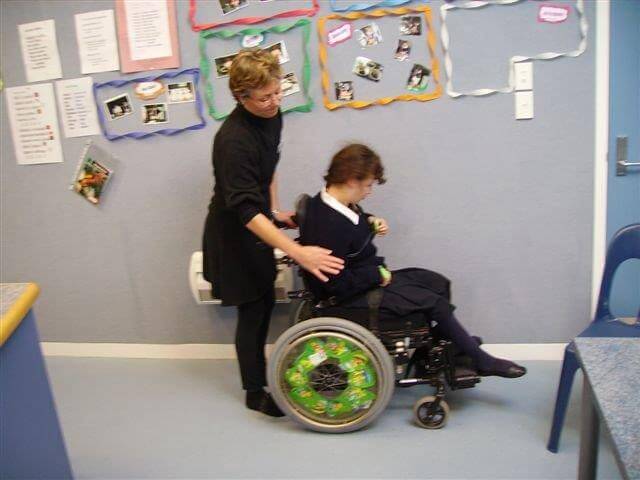 A learner is sitting in wheelchair with her supporter standing behind her touching her right arm.