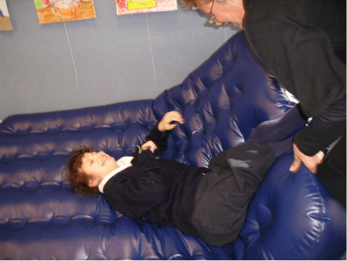 Sam is lying on their airbed pushing the O & M Specialist away with the legs.