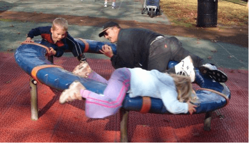Two children and their dad laying on their fronts spinning on a playground carousel.