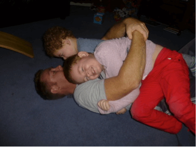 Two children laying on top of their dad on the floor playing, rough and tumble.