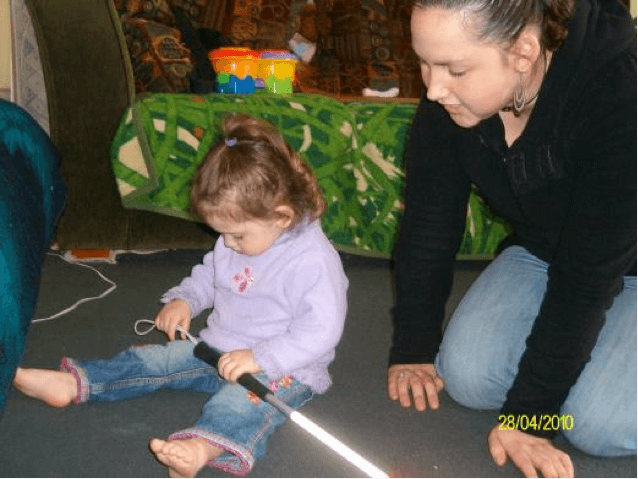 Young child sitting on the floor holding the top of a cane with adult sitting to her left watching on.