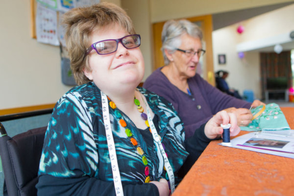 Katelyn sits at a table with her carer who is stitching