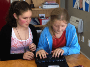 Two students sitting at a table, one has hands resting on BrailleNote
