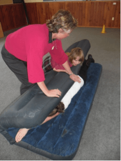 A child and adult pushing on another person rolled in an airbed