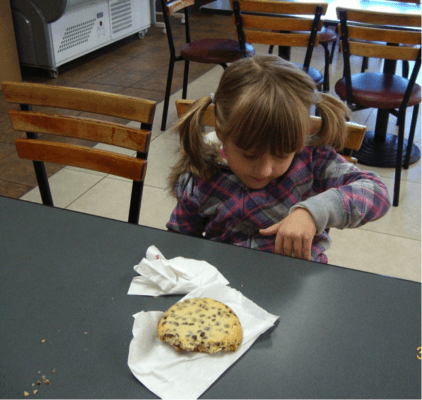 Child sitting at a table in a food court with a biscuit on the table, her left hand resting against the edge of the table