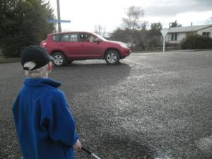 Child standing on the side of the road, holding his cane, watching and listening as a red car moves past him