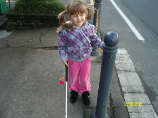 Child standing to the right of bollard beside a road crossing with cane in her right hand, trailing the chain that connects the bollards