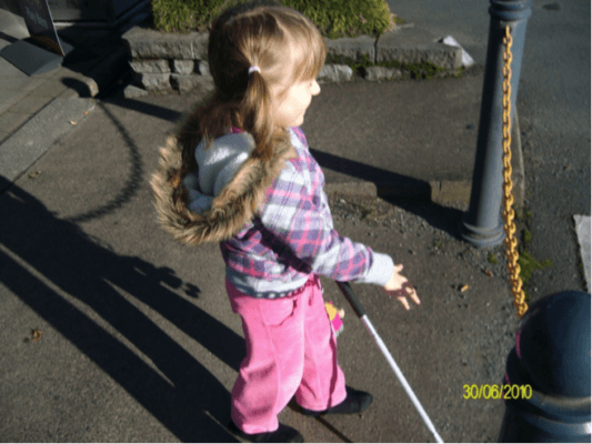 Child walking toward road crossing with cane in her left hand and right hand out in front of her