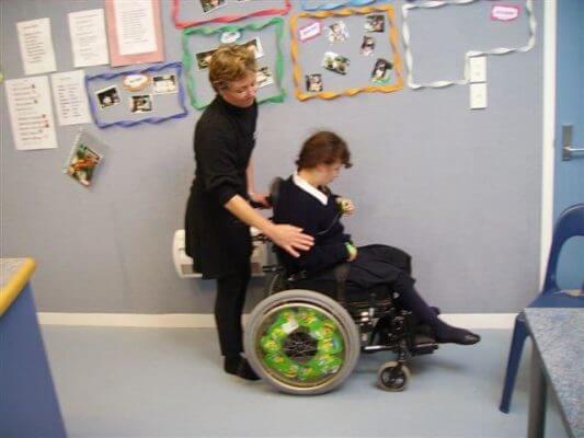A learner is sitting in wheelchair with her supporter standing behind her touching her right arm