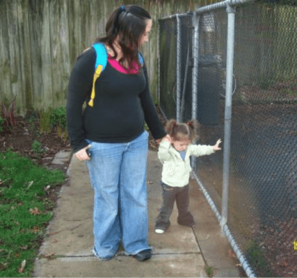 Mum with her toddler walking along a fence. Mum is guiding toddler, toddler is trailing hand along the fence
