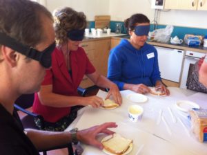 Three blindfolded adults sitting at a table buttering bread