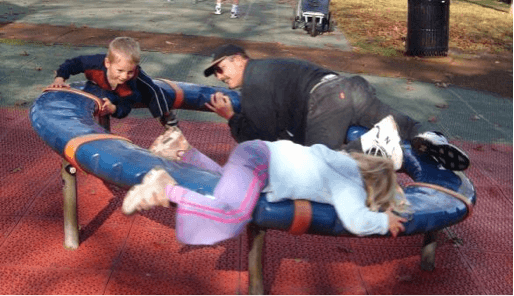 Two children and their dad laying on their fronts spinning on a playground carousel