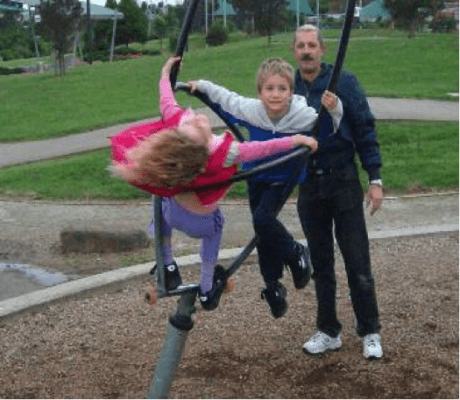 Two children on a rotating peice of playground equipment being watched by an adult