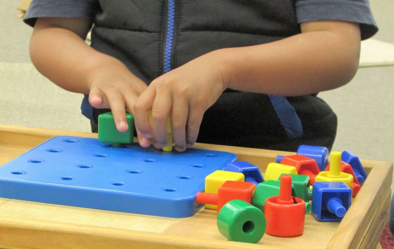 A boy who is blind is feeling the holes in the pegboard and is placing the pegs into the board