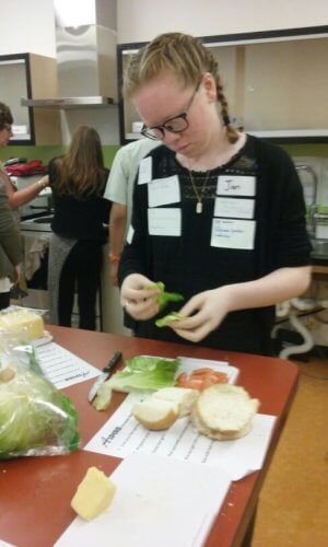 Female student stands at kitchen bench preparing salad items to go into burger