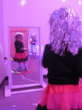 Aisha standing looking in the mirror, her reflection shown to the camera