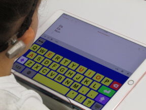 Ela sits in front of a table with an iPad. Keedogo Plus keyboard with large coloured keys is on the lower screen, Ela Ela Mum typed on top half