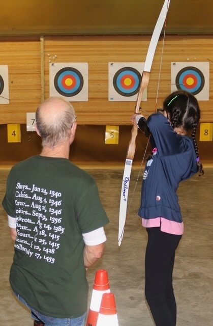 A student is holding a archery bow and arrow and aiming for a bullseye target with her instructor looking on beside her