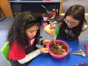 2 students are sitting at a table stirring a baking mixture that is in a bowl. 1 student is holding the bowl and the other student is holding a wooden spoon in her hand to do the stirring.