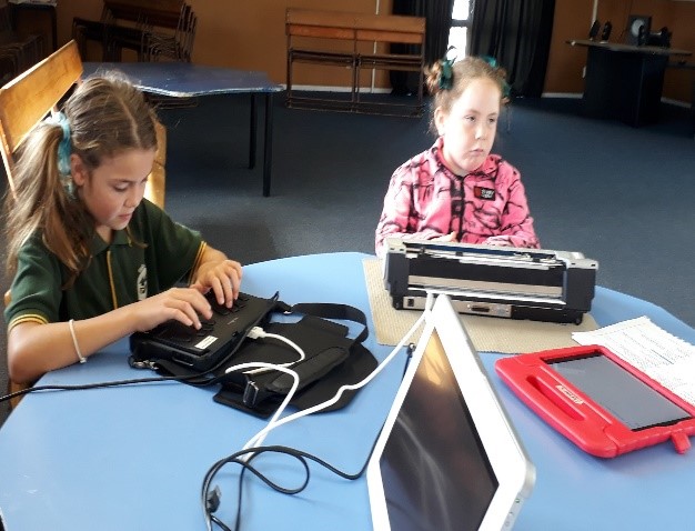 2 students sitting a a table with various pieces of technology and using their Braille devices to write letters and emails