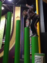A student is balancing on a tall green cylinder at a climbing and jumping centre. She is about to jump onto the ground.