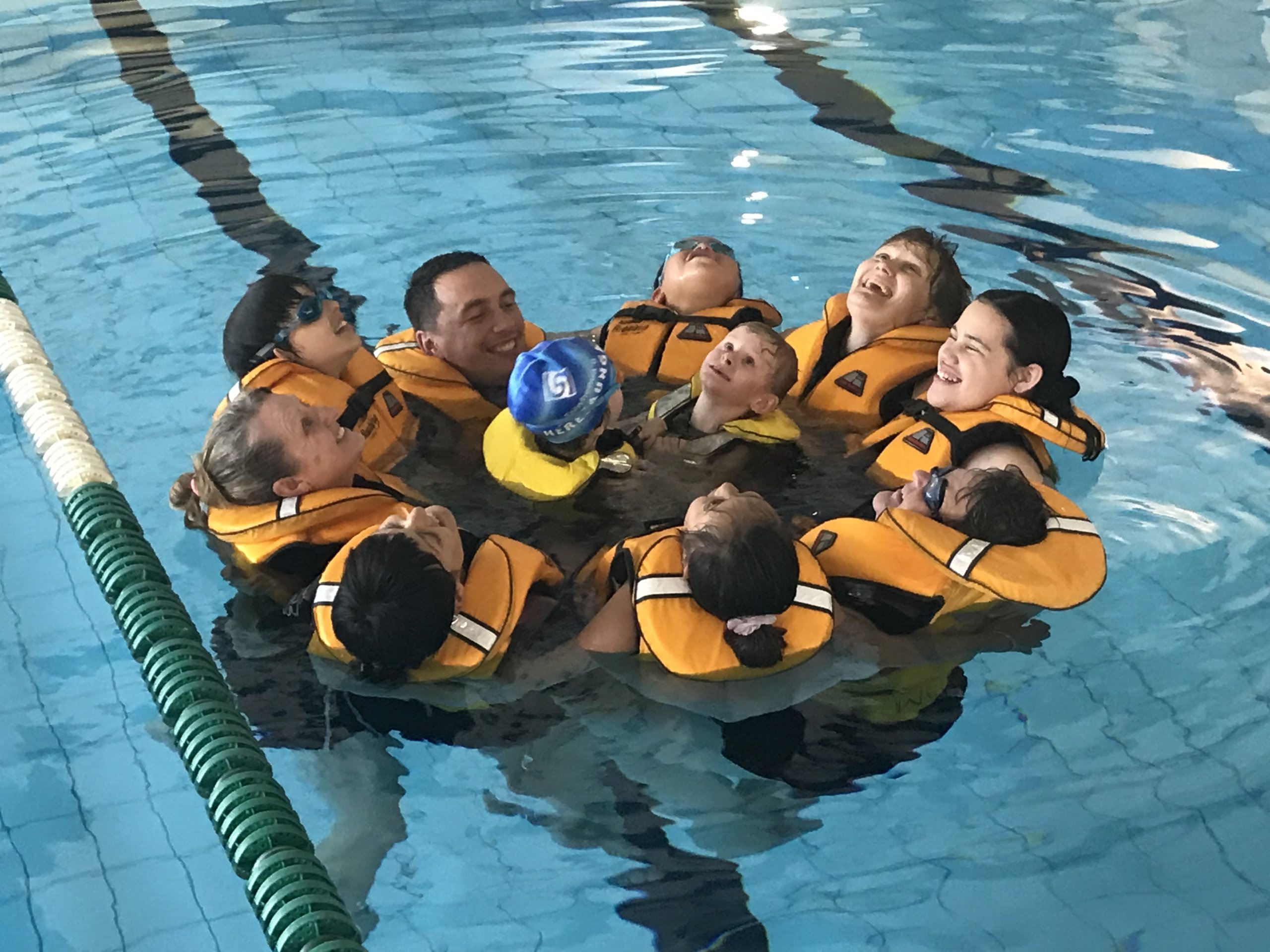 8 Ākonga and their instructors are all wearing lifejackets and are joined together huddled in a circle in the swimming pool