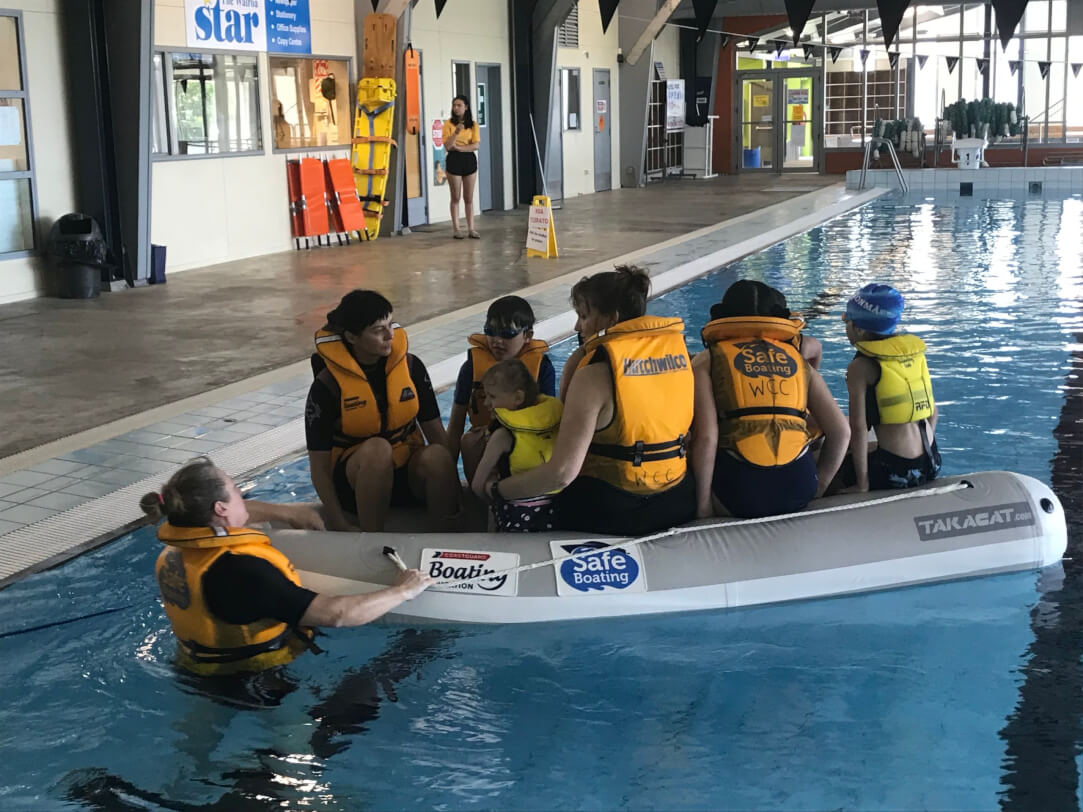 8 Ākonga wearing life jackets in an inflatable raft floating in an indoor swimming pool are getting ready to jump out vackwards with support from thier AUSTSwim instructor who is holding onto the side of the raft