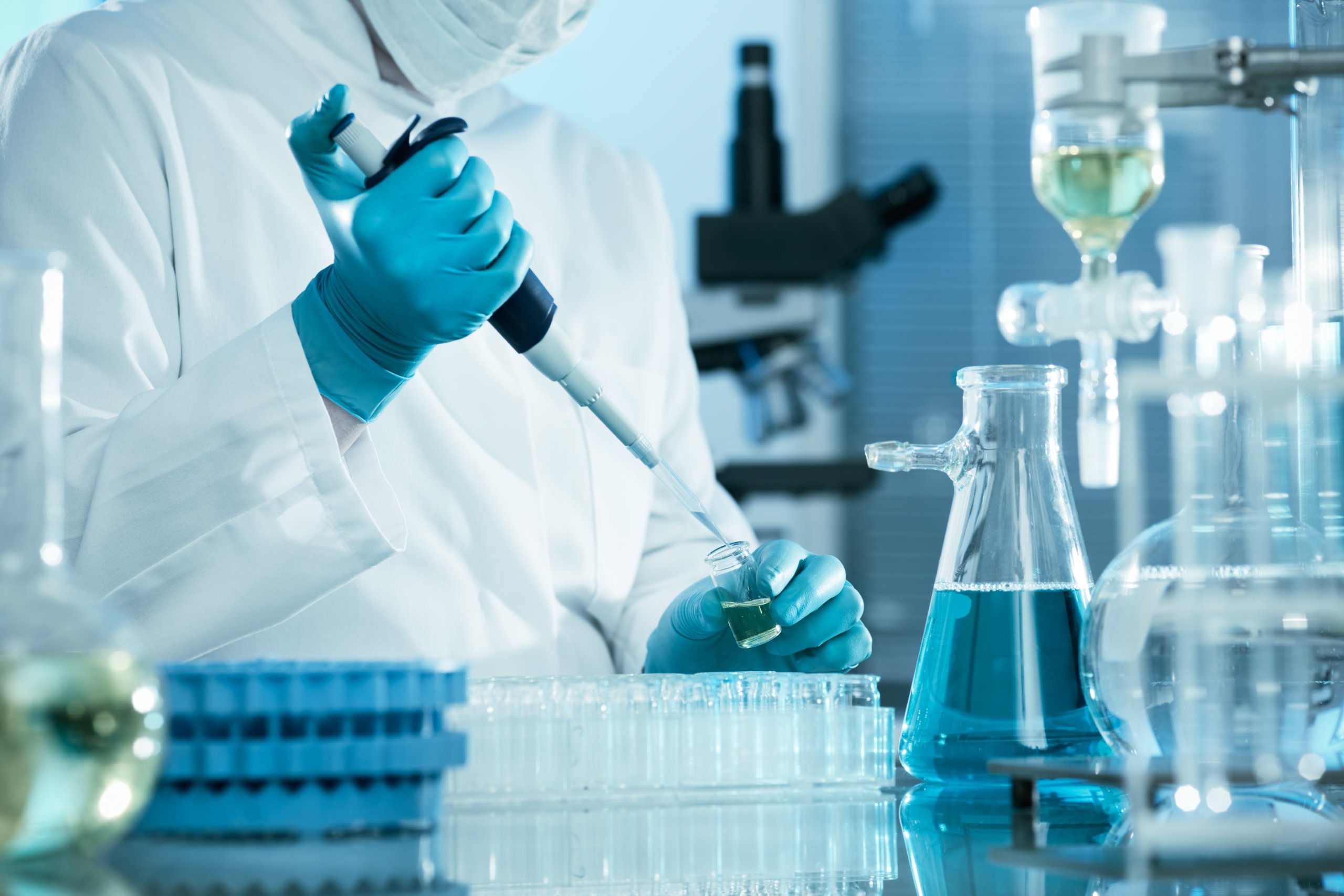 Science technician in white coat holding beaker surrounded by vials with blue liquid