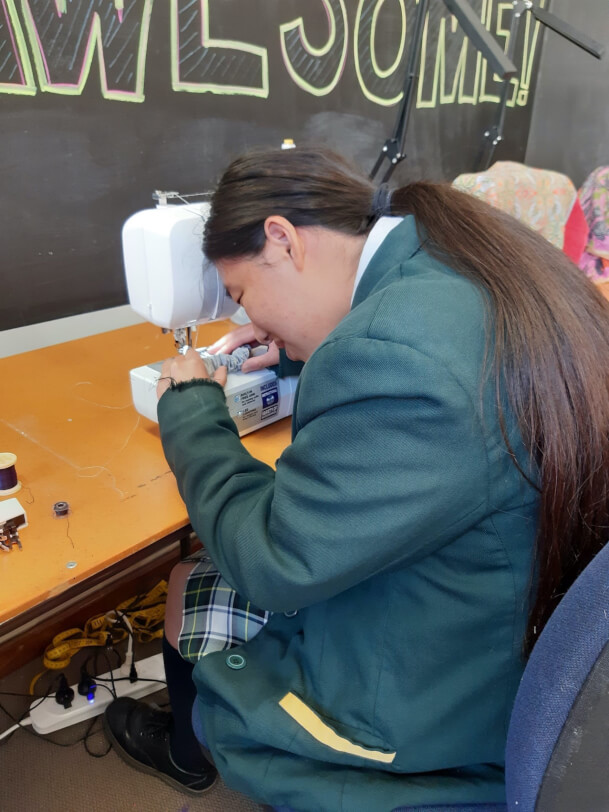 Figure 2 - A student is sitting at a sewing table with her hands guiding the needle into her fabricsewing machine `