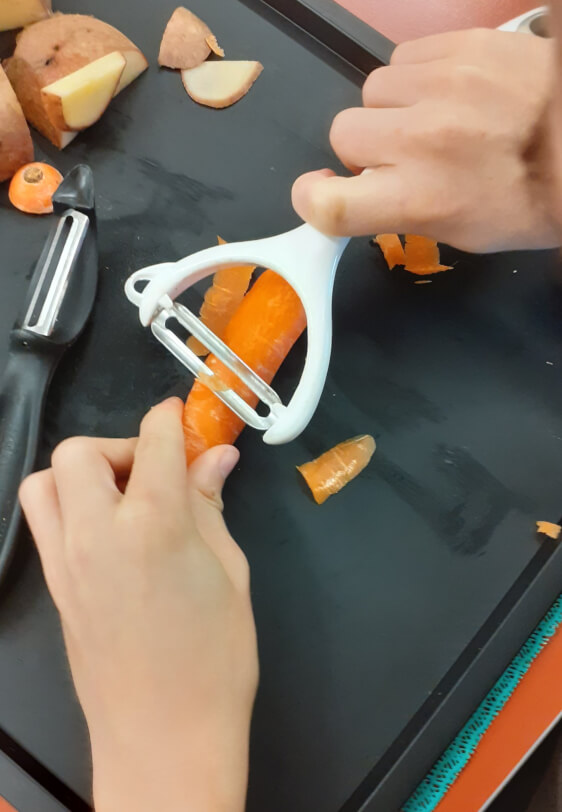 Student is holding a carrot against the surface of a black tray with his left hand, and using a bigger grip handled white peeler in his right hand to peel it using a scrape to the right.