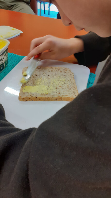 Joshua is using his right hand to spread margarine on to a slice of bread with a bigger grip handled knife. He is using a sandwich spreading board with non-slip matting underneath.