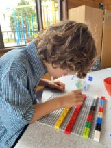 A child is sitting in front of a grey LEGO base plate which has four rows of LEGO attached. The top two rows are coloured LEGO, the two lower rows are LEGO® Braille Bricks. One LEGO® Braille Bricks row is red and has one top left dot on the brick that represents the letter A and number 1. The second LEGO® Braille Bricks row is yellow and has the top and middle left dots on the brick which represents the letter B and number 2
