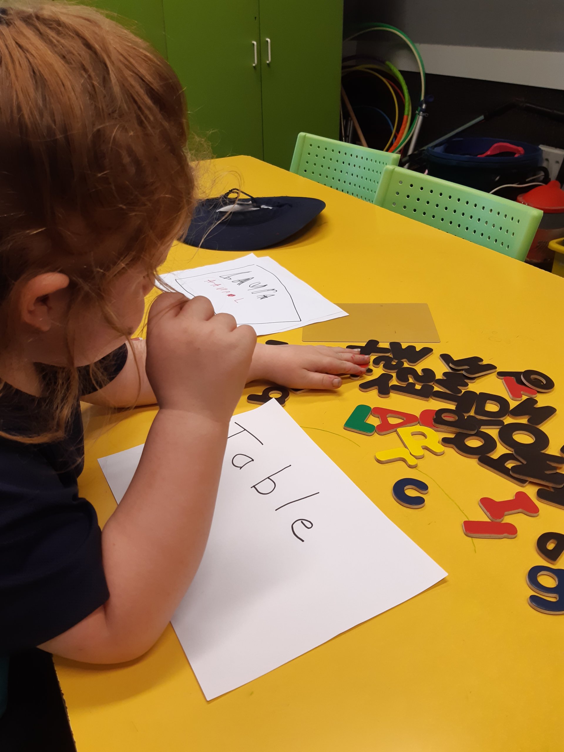 Child is sitting at a table looking at some wooden letters to match to a word on a piece of paper
