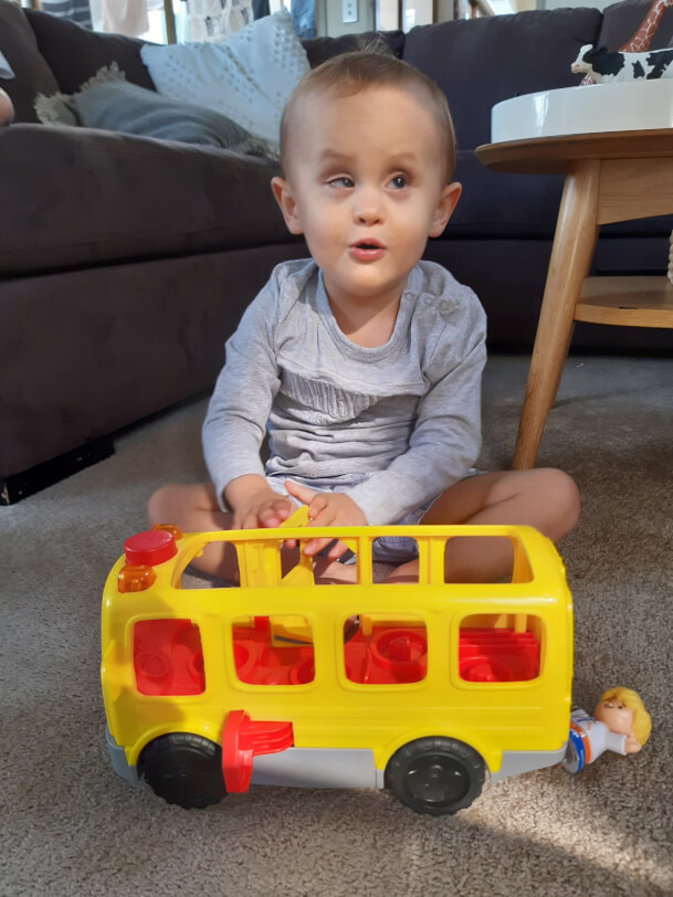 Figure 1 - Tobi sits on the floor behind a large yellow bus toy. He smiles and holds a part of the bus toy in his hands. 