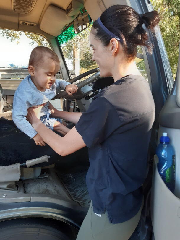 Figure 2 - Tobi's mum holds him while sitting in the driver's seat of the bus. He has one hand on the seat and one hand on the steering wheel. Hannah looks at Tobi and smiles, Tobi smiles. 